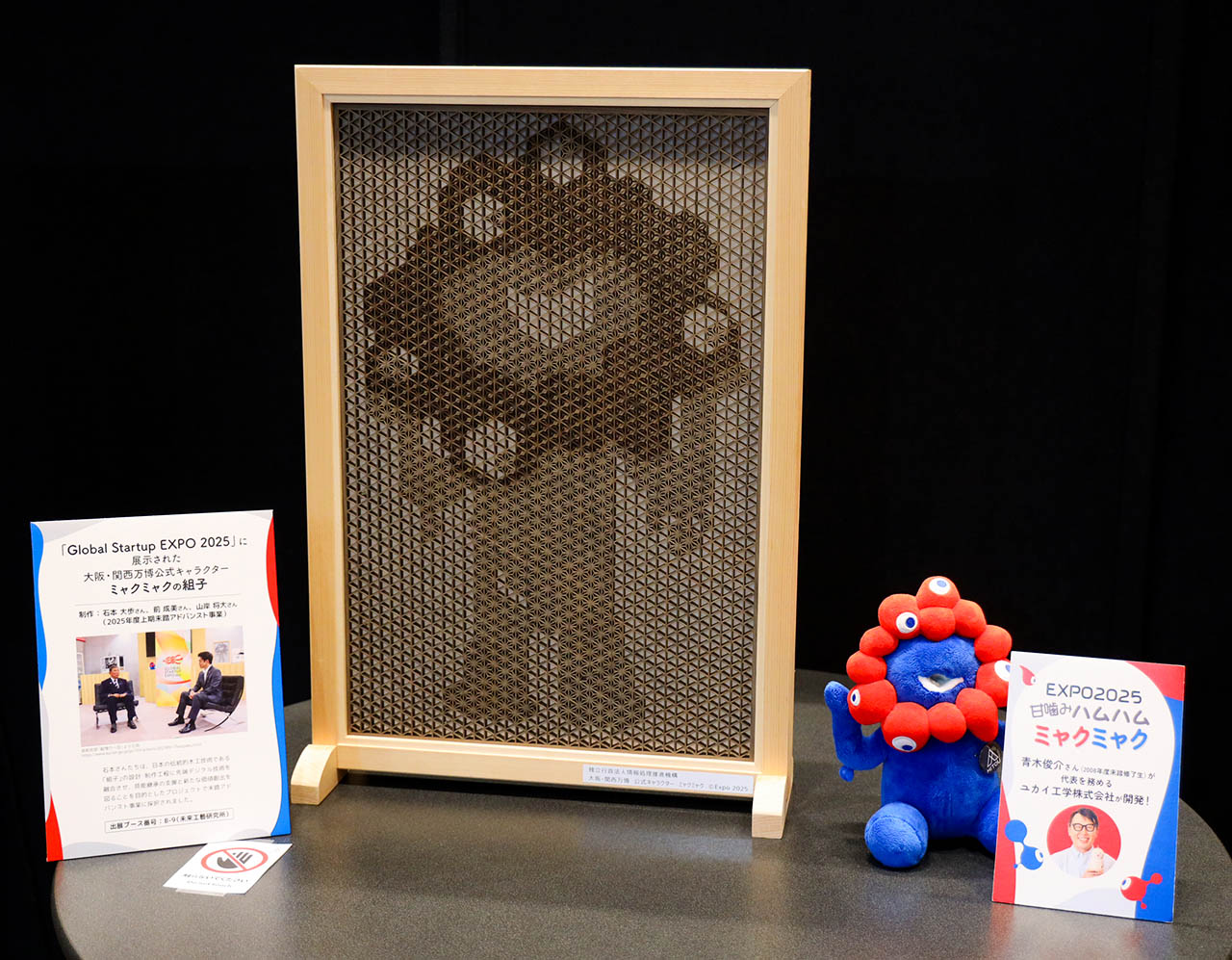 左が組子細工でミャクミャクの姿を表した木枠パネル、右がぬいぐるみ「甘噛みハムハム ミャクミャク」とそれぞれの説明パネルの展示